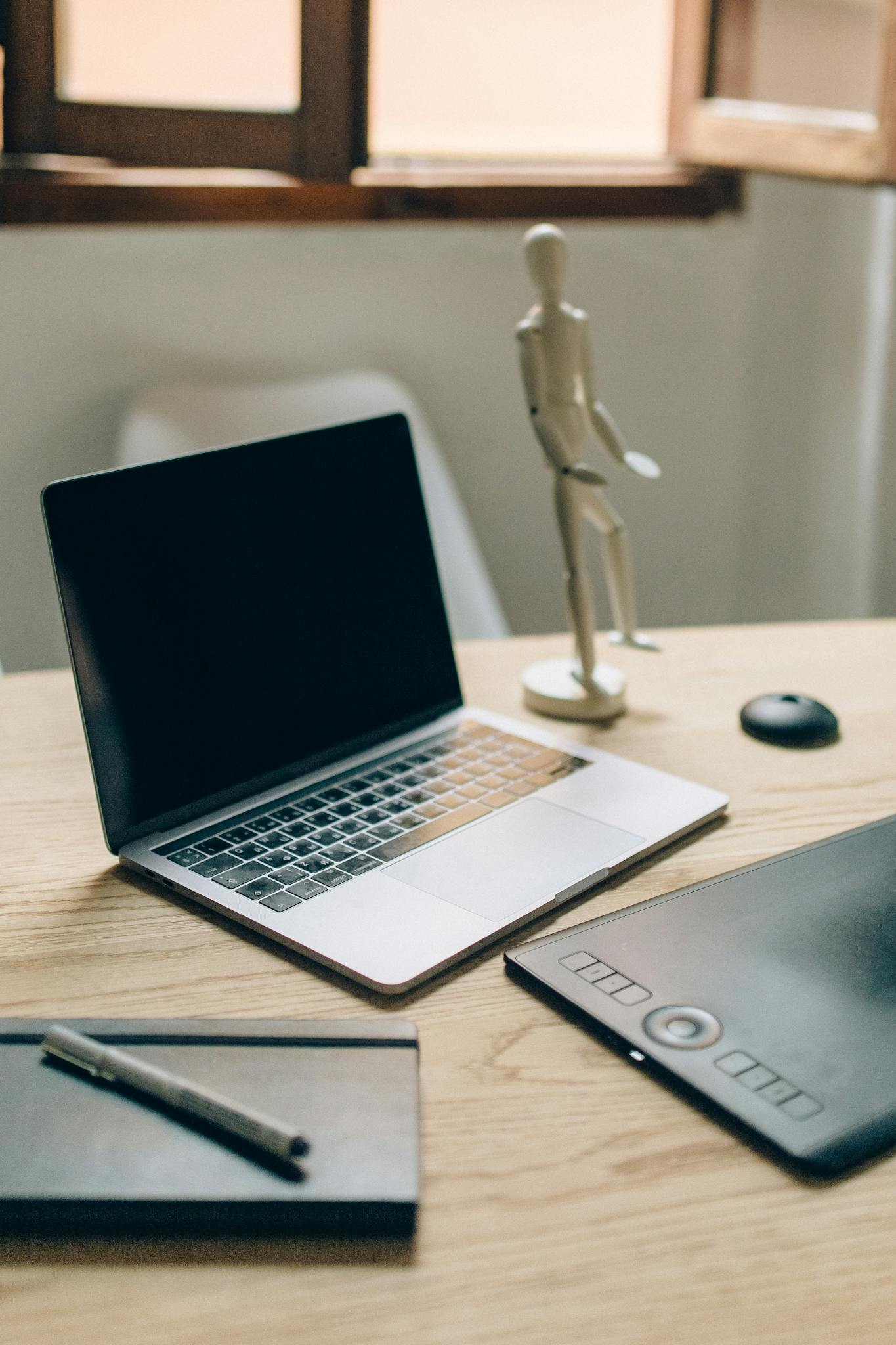 A minimalist workspace featuring a laptop, sketchpad, and figurine on a wooden desk.